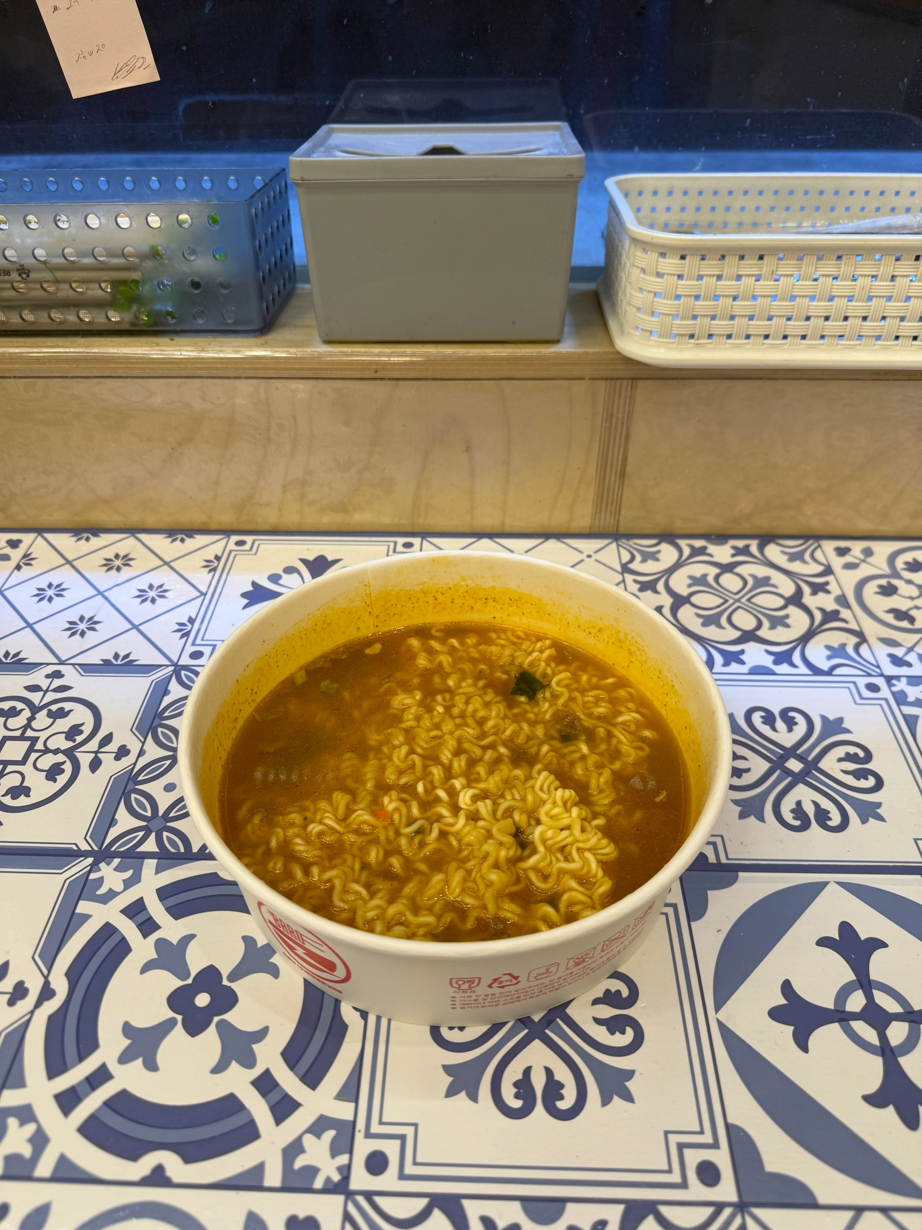 Auto-generated description: A bowl of instant ramen sits on a patterned table with various kitchen items in the background.