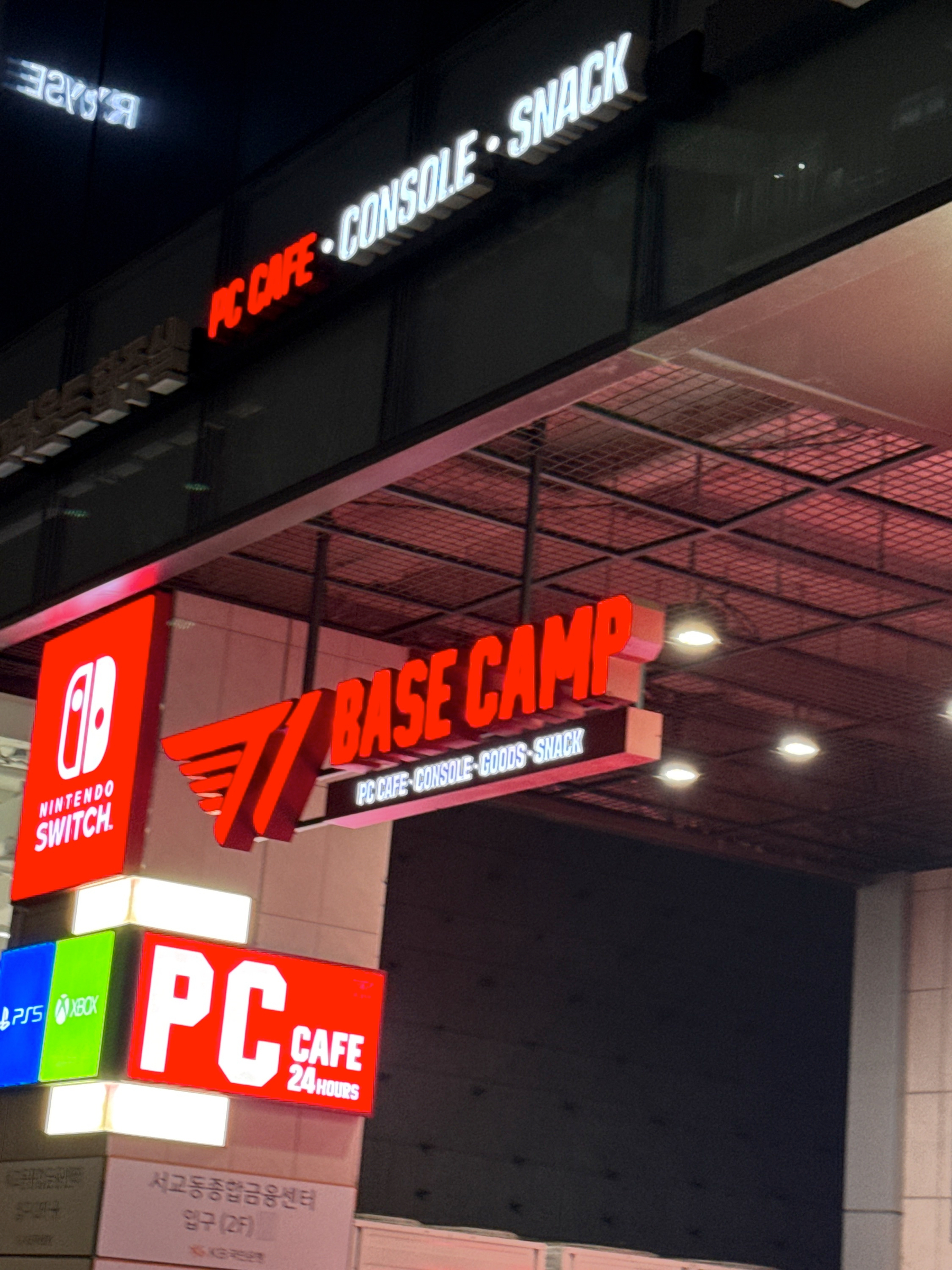 A storefront with illuminated signs features BASE CAMP alongside PC CAFE · CONSOLE · GOODS · SNACK and logos for Nintendo Switch and Xbox.