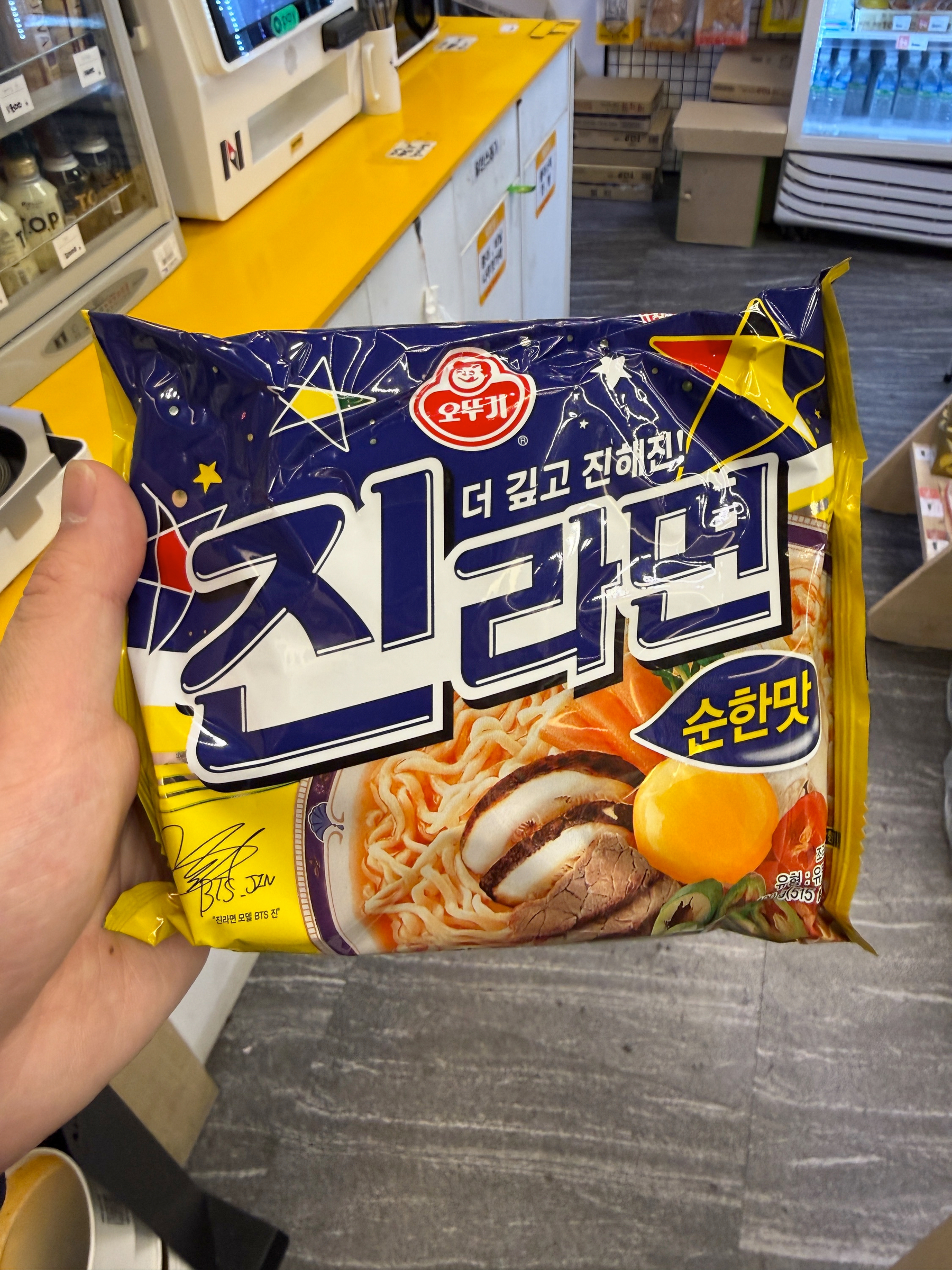 Auto-generated description: A person is holding a package of instant noodles with Korean labeling, depicting ingredients like meat and eggs, inside a store.