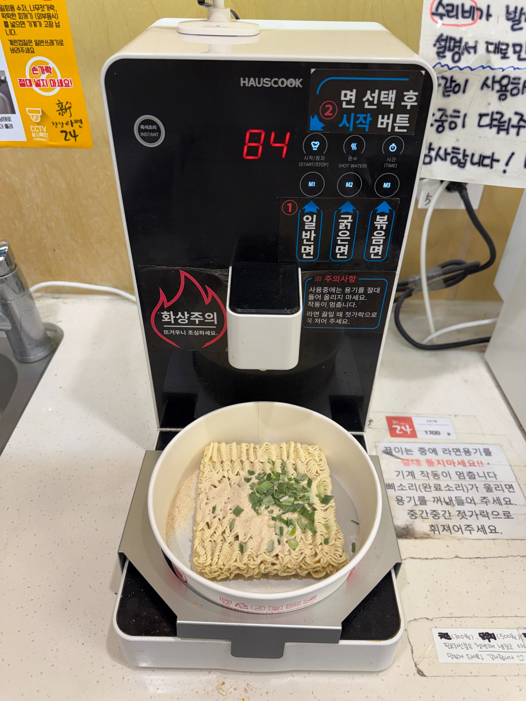 Auto-generated description: A noodle cup is being prepared under a hot water dispenser, with instant noodles and seasoning visible.