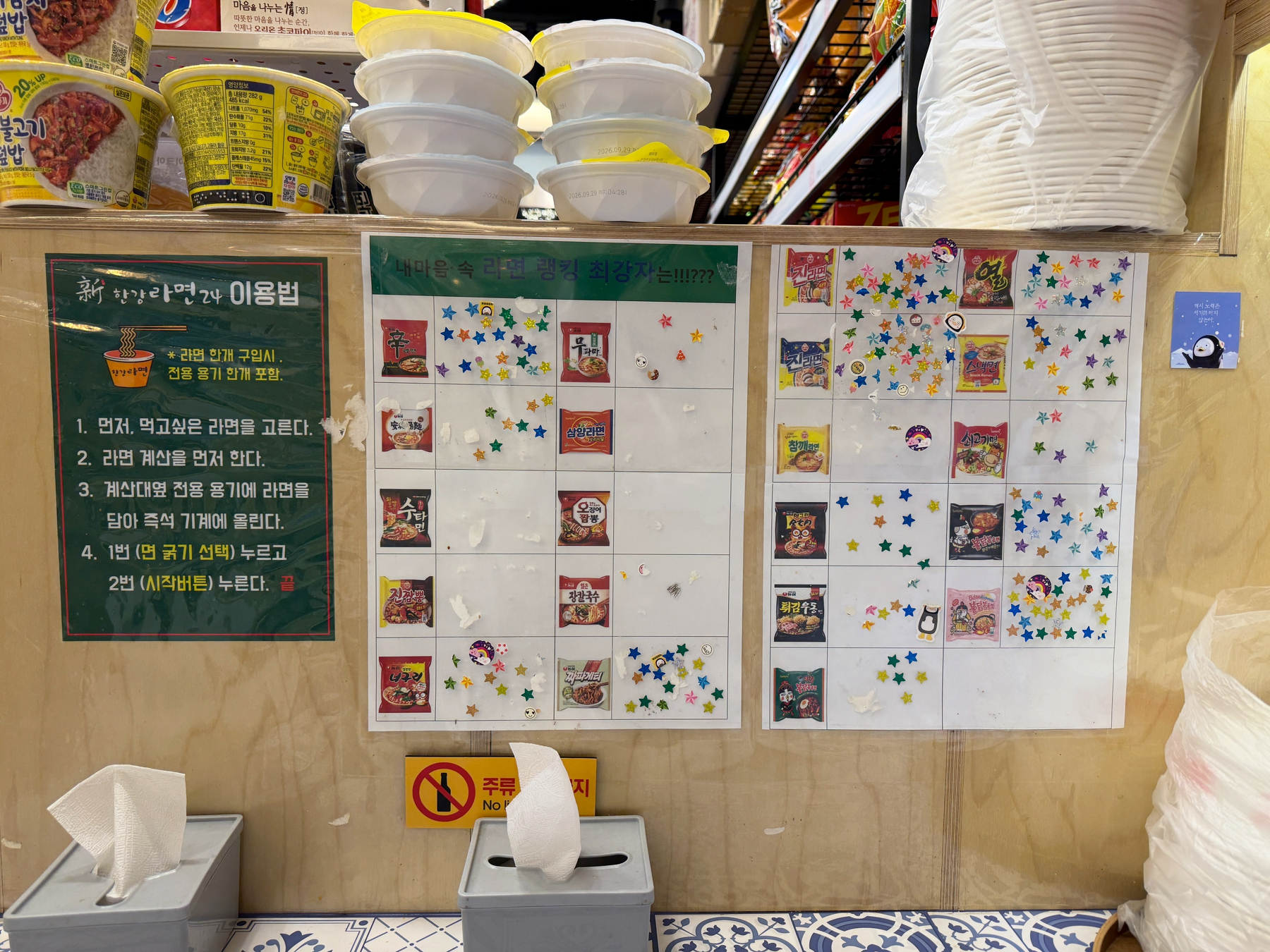 Auto-generated description: A display features a wall menu with images of ramen varieties and numerous colorful star stickers, along with shelves of bowls and containers above.