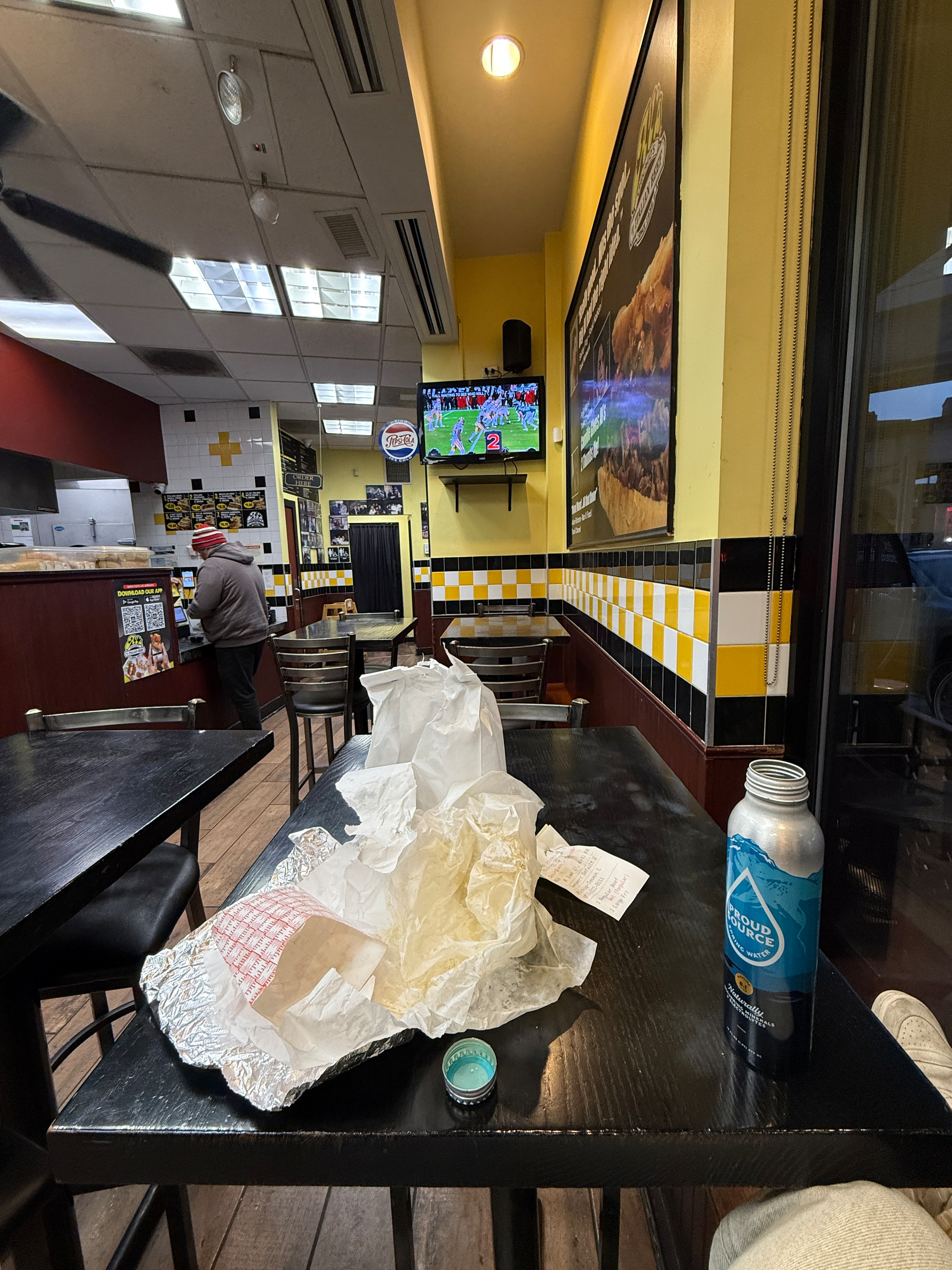 A casual restaurant interior features tables, discarded food wrappers, a beverage, and a television displaying a sporting event.