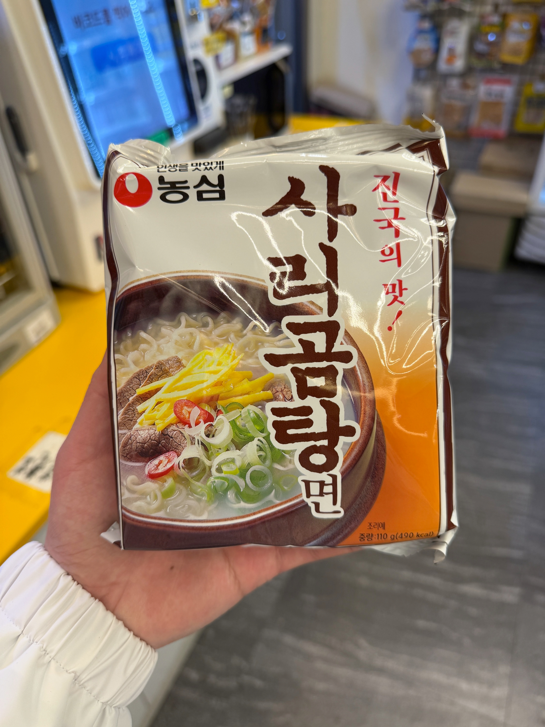 Auto-generated description: A person is holding a pack of instant noodles featuring a bowl of broth with green onions and beef on the packaging.