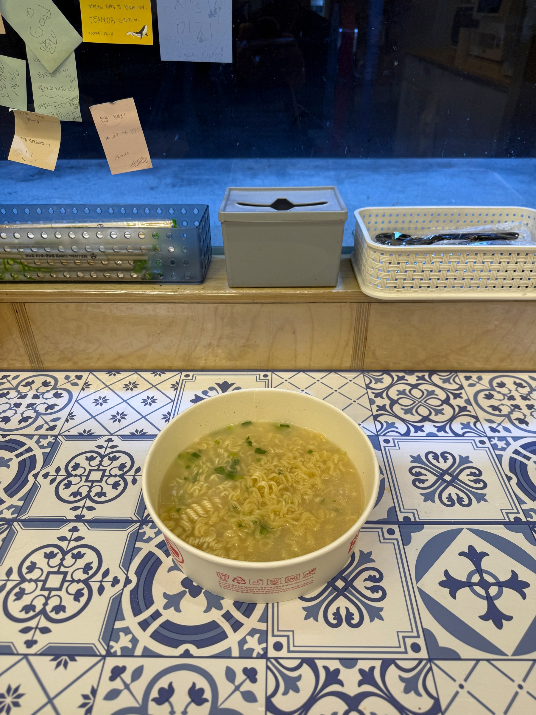 Auto-generated description: A bowl of noodles sits on a decorative blue and white tiled table, with notes and containers visible in the background.