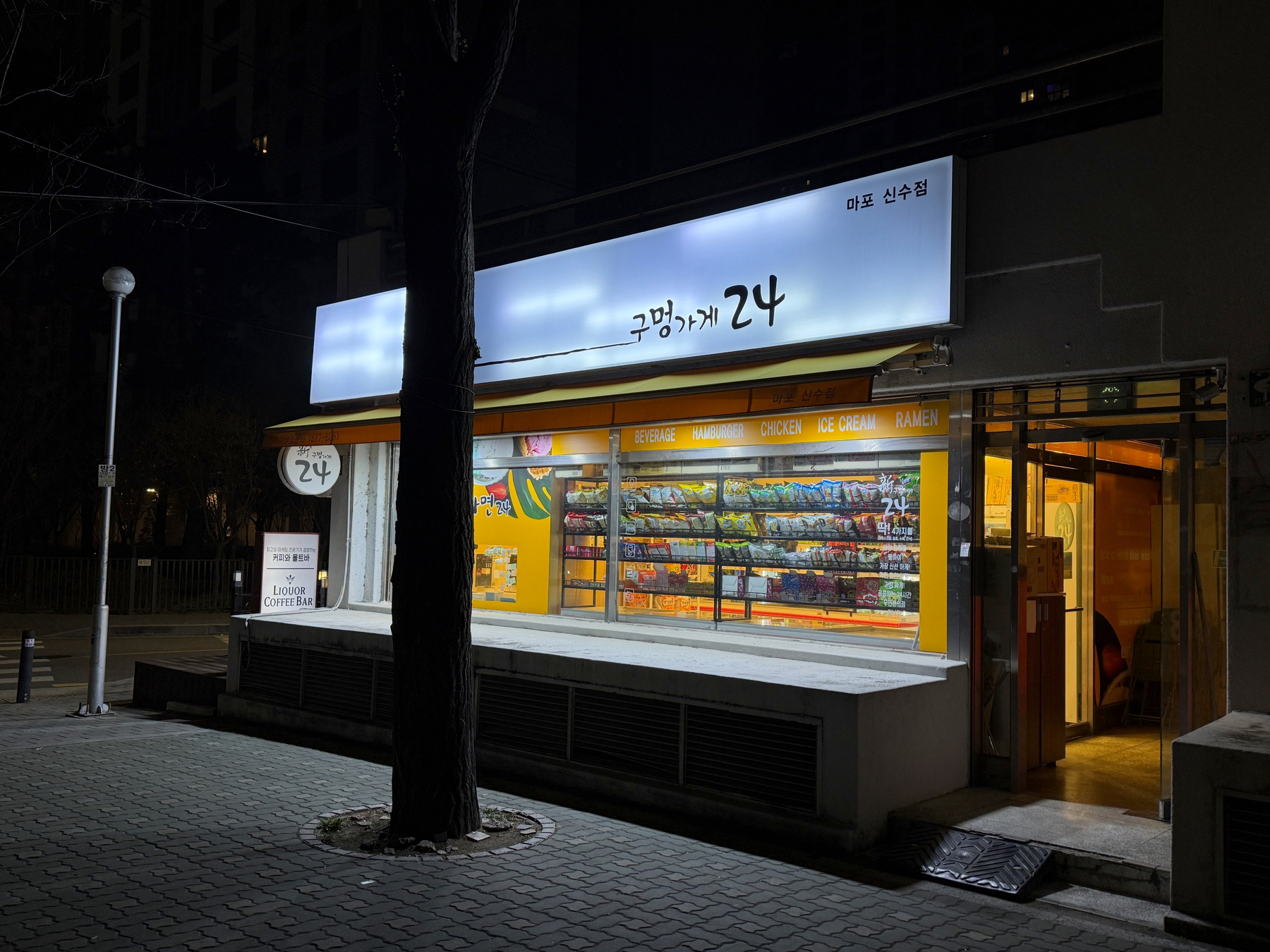 Auto-generated description: A convenience store with a brightly lit interior and colorful products is situated on a quiet street at night.